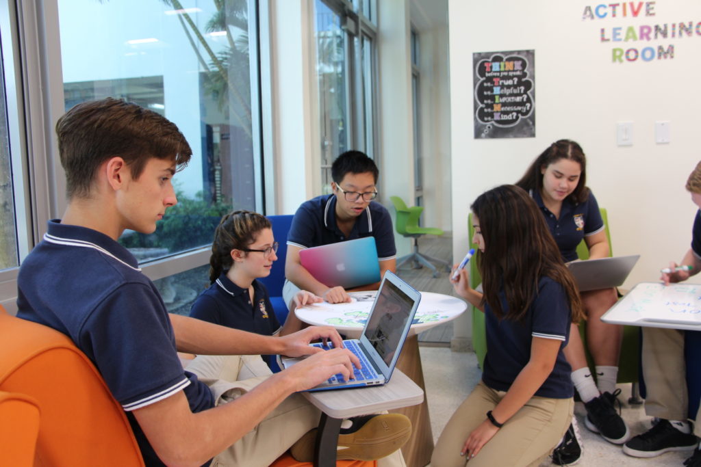 A group of middle and upper school students sit in the library space with their laptops and white boards to work on a group project.