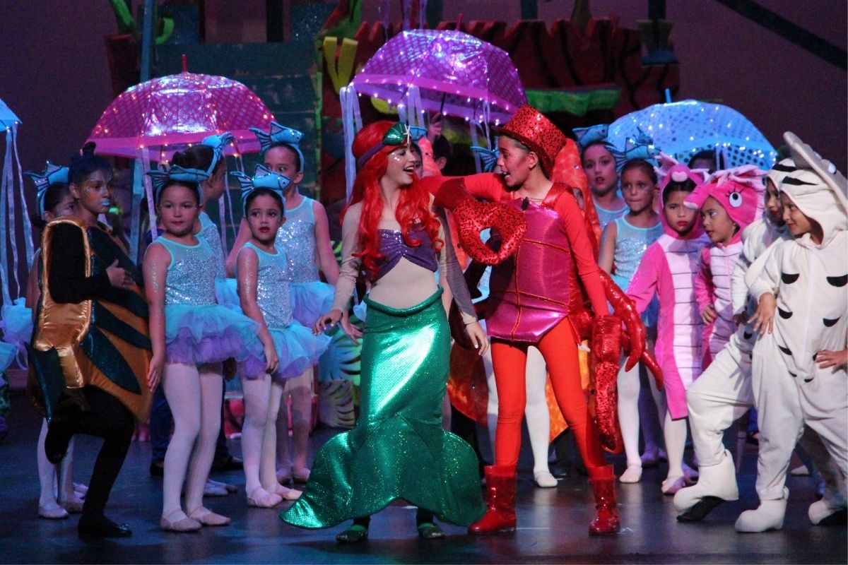 Students playing Ariel, in her full mermaid costume, and Sebastian, in their full red crab costume, stand center stage. Flounder the fish in blue and yellow costume stands in the back with the other students playing sea creatures.