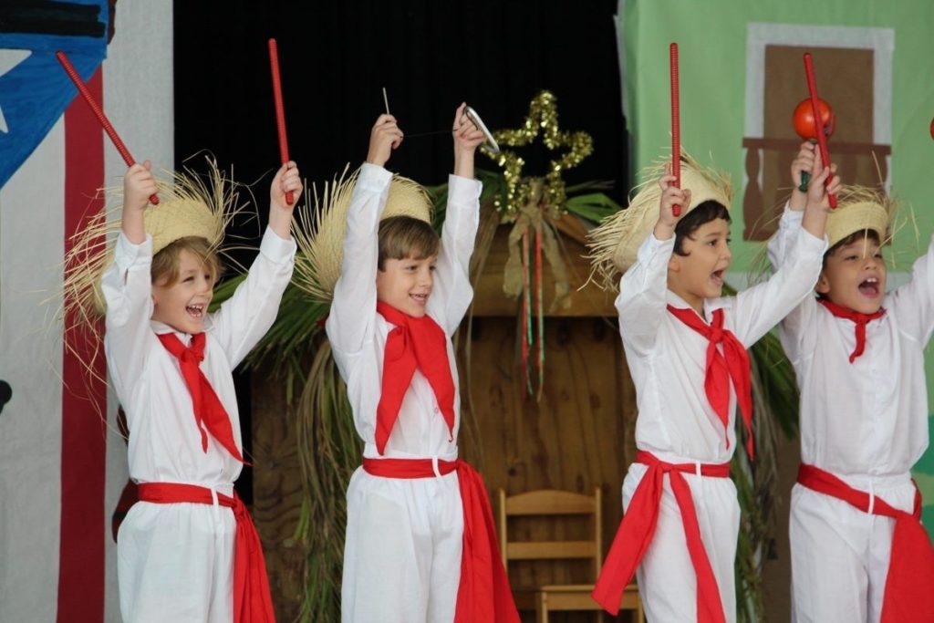 Four young male Robinson students wearing jibaro costumes with red neck ties and waist bands, holding up red sticks, red maracas and a silver triangle.