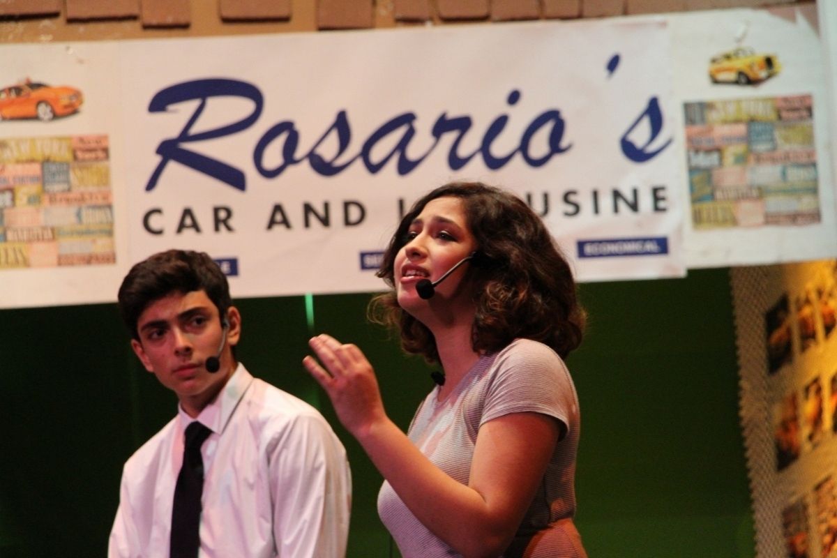 A young boy in a white dress shirt and black tie looks at a girl who is singing and looking into the audience, both wearing microphones. There is a sign behind them that states 'Rosario's car and limousine'.