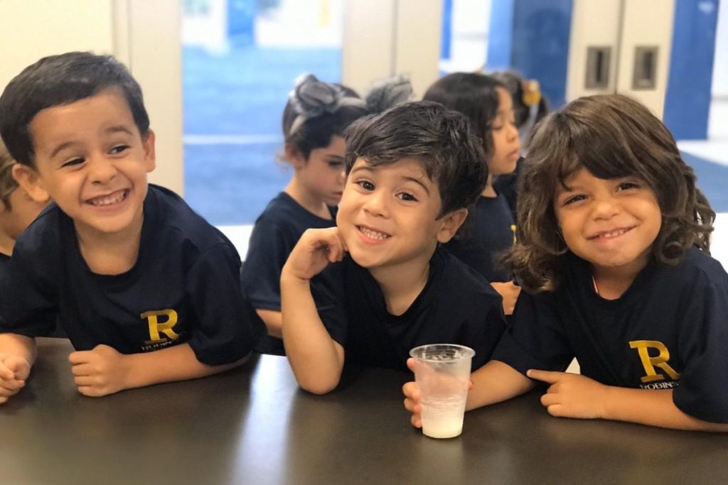Three male preschool Robinson students smile while sitting next to each other, the one of the far right holding a glass of milk.