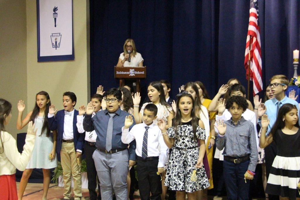 A group of Elementary School students in professional dress wear hold their right hand up while initiating into the Elementary National Honor Society, with Head of School Cindy Ogg behind them.