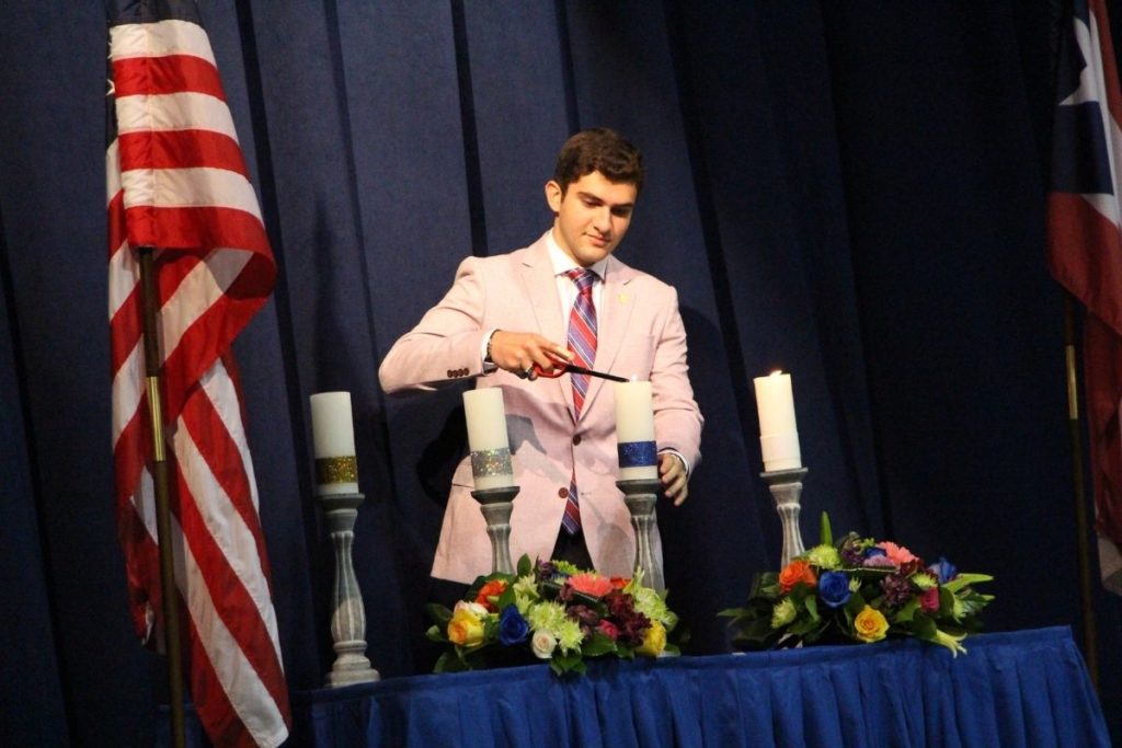 A male student in a pink suit lights a candle at Robinson School's National Honor Society installation.