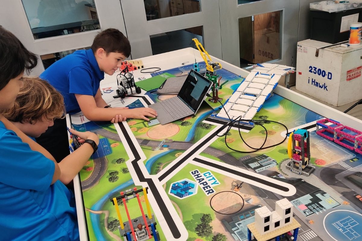 Three male Robinson School students in royal blue shirts program their robot on top of a map specifically for the robot to run on.