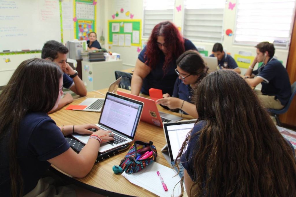 A teacher leans over to help a student with her online course on her laptop, with her classmates sitting at the same table taking their own virtual courses on their personal laptops.