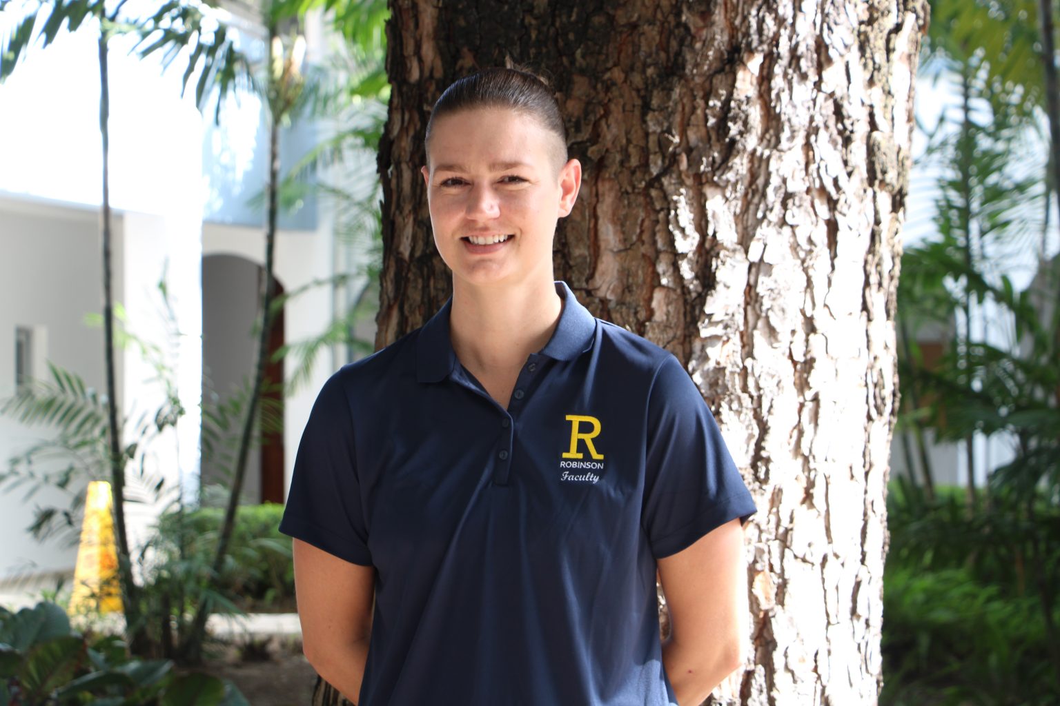 Elementary School Dean of Students Shaina Sullivan stands while wearing a navy blue polo shirt with the yellow Robinson R on the right hand side and with a tree behind her.